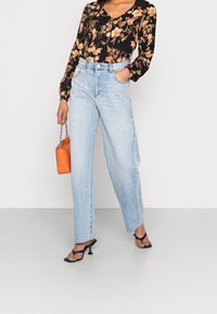 Blusa nera floreale con maniche lunghe, jeans a vita alta blu chiaro, décolleté neri con tacco a spillo e una borsa arancione.