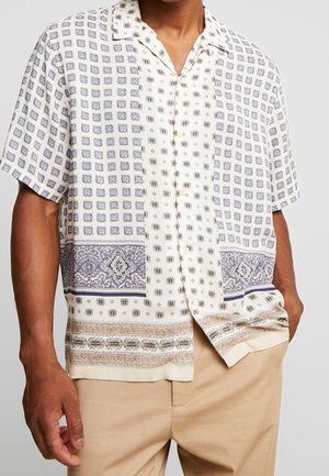 Homme portant une chemise blanche à manches courtes avec des motifs géométriques et cachemire beiges et gris, associée à un pantalon beige clair.