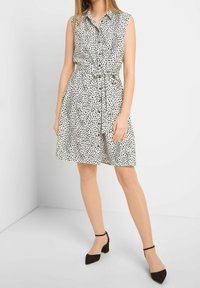 Robe sans manches blanche à pois noirs, boutonnée sur le devant, ceinture nouée à la taille et jupe évasée. Associée à des chaussures à talons avec bride à la cheville noires.