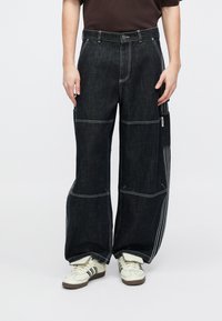 Schwarze Jeanshose mit weitem Bein, kontrastierende weiße Nähte und eine Seitentasche. Das Material hat eine strukturierte Oberfläche.