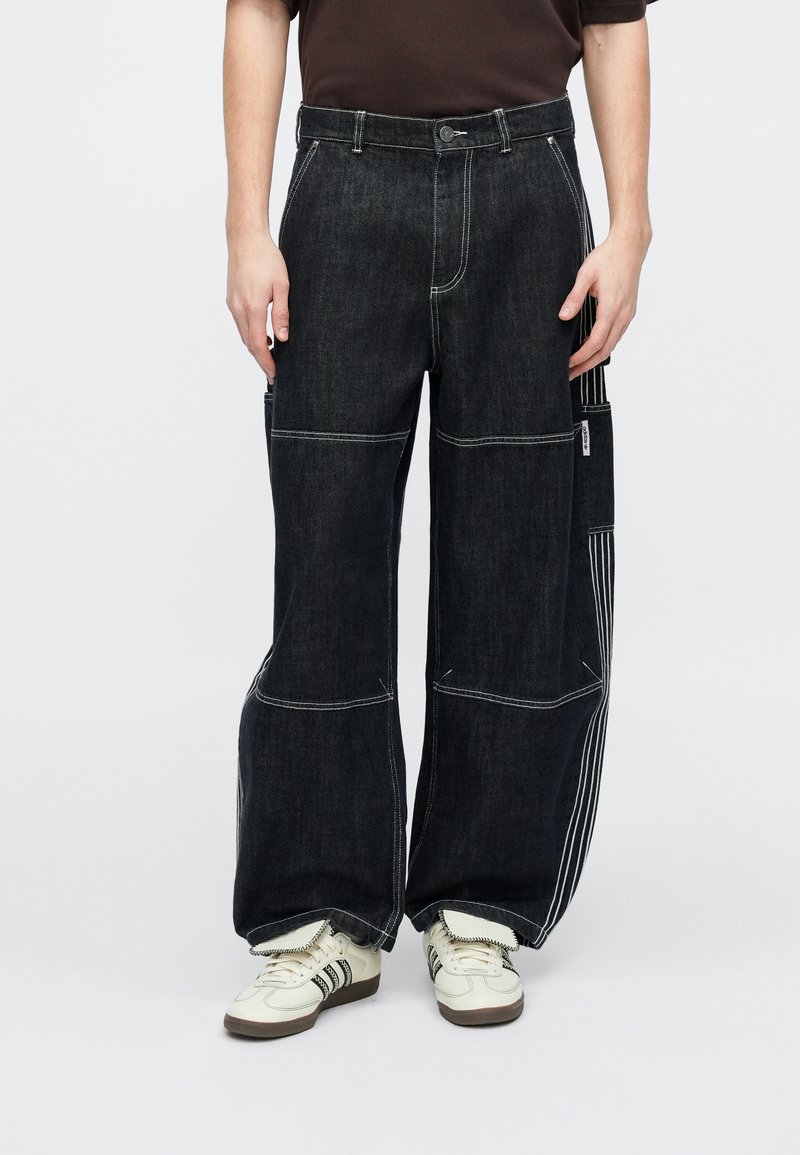 Schwarze Jeanshose mit weitem Bein, kontrastierende weiße Nähte und eine Seitentasche. Das Material hat eine strukturierte Oberfläche.