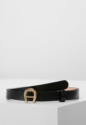 Ceinture en cuir noir avec une boucle dorée en forme de fer à cheval, enroulée et présentée sur une surface blanche sur un fond gris clair.