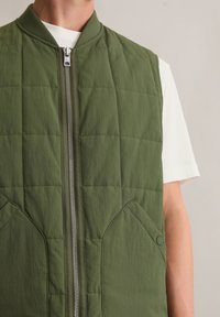 Pikowana zielona kamizelka z zamkiem błyskawicznym z przodu, okrągłym dekoltem i bocznymi kieszeniami. Wykonana z lekkiego materiału o matowym wykończeniu.