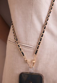 Zelta ķēde ar melniem akcentiem, pievienota caurspīdīgam turētājam, guļot uz bēša auduma. Ķēdei ir aizdares klips drošai stiprināšanai.