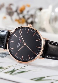 Reloj analógico negro con manecillas y marcadores en tono dorado, un diseño minimalista y una correa de cuero negro con costuras blancas.