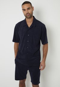 Camicia a maniche corte in terry blu navy con bottoni sul davanti, abbinata a pantaloni corti coordinati. Tessuto morbido e vestibilità rilassata.