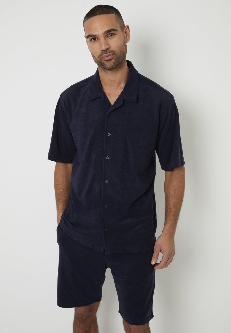 Camicia a maniche corte in terry blu navy con bottoni sul davanti, abbinata a pantaloni corti coordinati. Tessuto morbido e vestibilità rilassata.