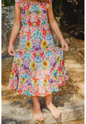 Jeune enfant debout pieds nus à l'extérieur, portant une robe fleurie colorée, tenant l'ourlet des deux mains.