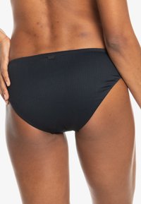 Black ribbed bikini bottom with a mid-rise design, featuring a smooth texture and a subtle logo tag at the back for branding.