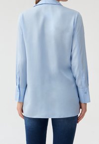 Camisa de manga larga azul claro con cuello, que presenta puños abotonados. Tela suave con un corte relajado, el dobladillo se sitúa por debajo de las caderas.