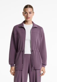 Lilla zip-up jakke med høj krave, snørre i taljen og frontlommer; blødt stof med en glat tekstur; parret med en hvid skjorte.