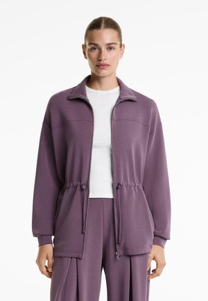 Purple zip-up jacket with a high collar, drawstring waist, and front pockets; soft fabric with a smooth texture; paired with a white shirt.