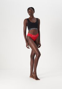 Μαύρο αμάνικο μπλουζάκι με λευκή λωρίδα με λογότυπο και κόκκινο μπικίνι με λευκό λογότυπο. Το ύφασμα φαίνεται λείο και εφαρμοστό, αναδεικνύοντας τη σιλουέτα του μοντέλου.