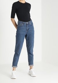 Černé fitted tričko, džíny s vysokým pasem z modrého denimu s ohrnutými manžetami, doplněné bílými teniskami. Obsahuje klasický design s pěti kapsami.