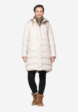 Woman wearing a light beige quilted winter coat, brown sweater, black leggings, and brown fur-lined winter boots standing on a white background.