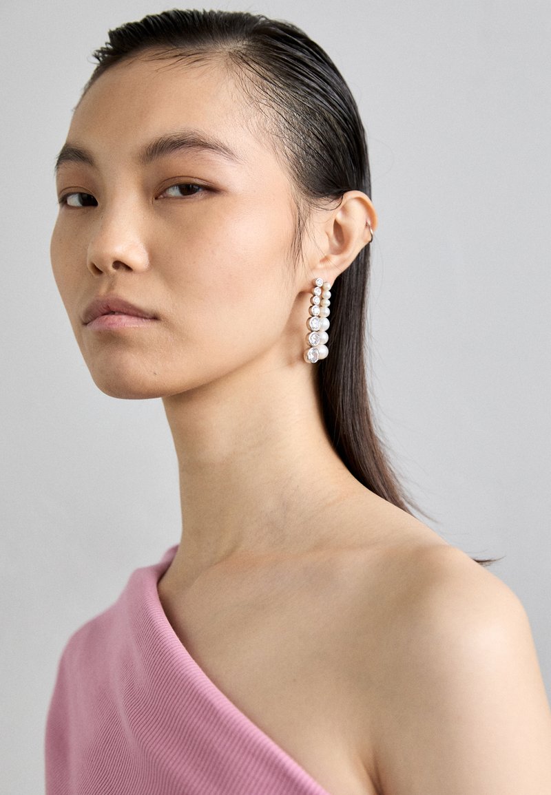 Διαμαντένια σκουλαρίκια δύο επιπέδων με εναλλασσόμενες σειρές μικρών μαργαριταριών και στρογγυλών διαφανών πετρών, κατασκευασμένα από ασημένιο χρώμα μέταλλο, φορεμένα από μια μοντέλο.