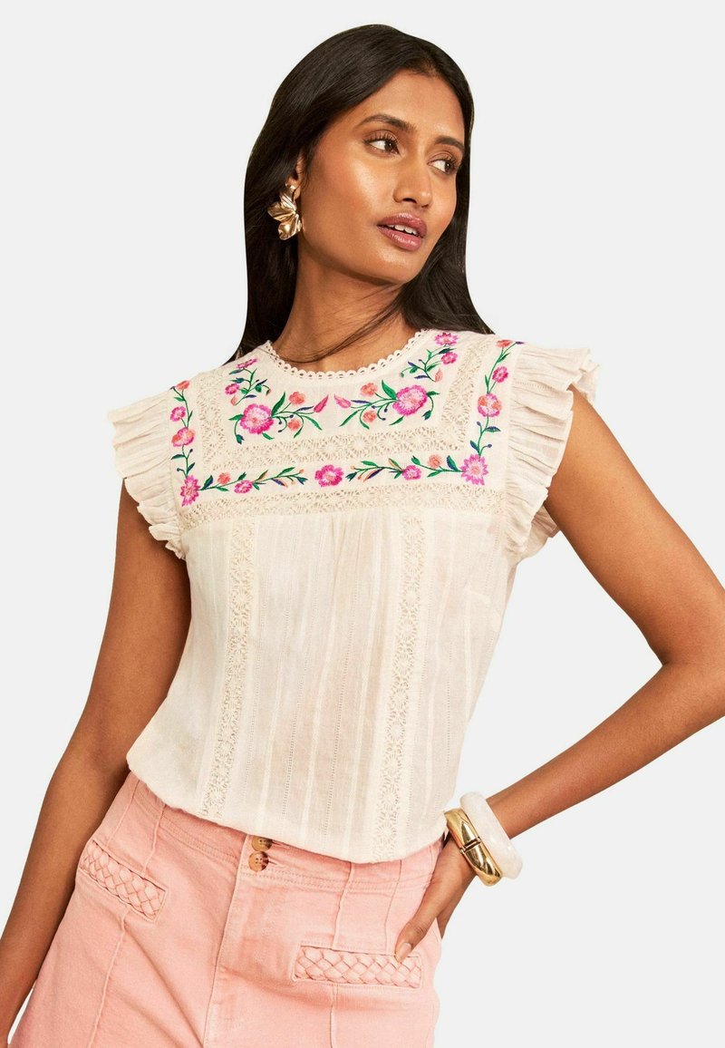 Woman with long dark hair wearing cream blouse with pink floral embroidery and ruffled sleeves, paired with light pink pants and gold jewelry.