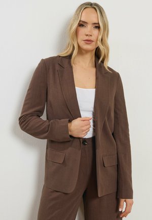 Blonde vrouw in een bruine colbert en broek met een wit topje, staand met één hand de kraag van haar colbert vasthoudend tegen een witte muur.