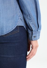 Primo piano di una mano di persona accanto alla tasca di un jeans blu scuro, indossando una camicia di denim a maniche lunghe azzurro chiaro con polsini abbottonati.