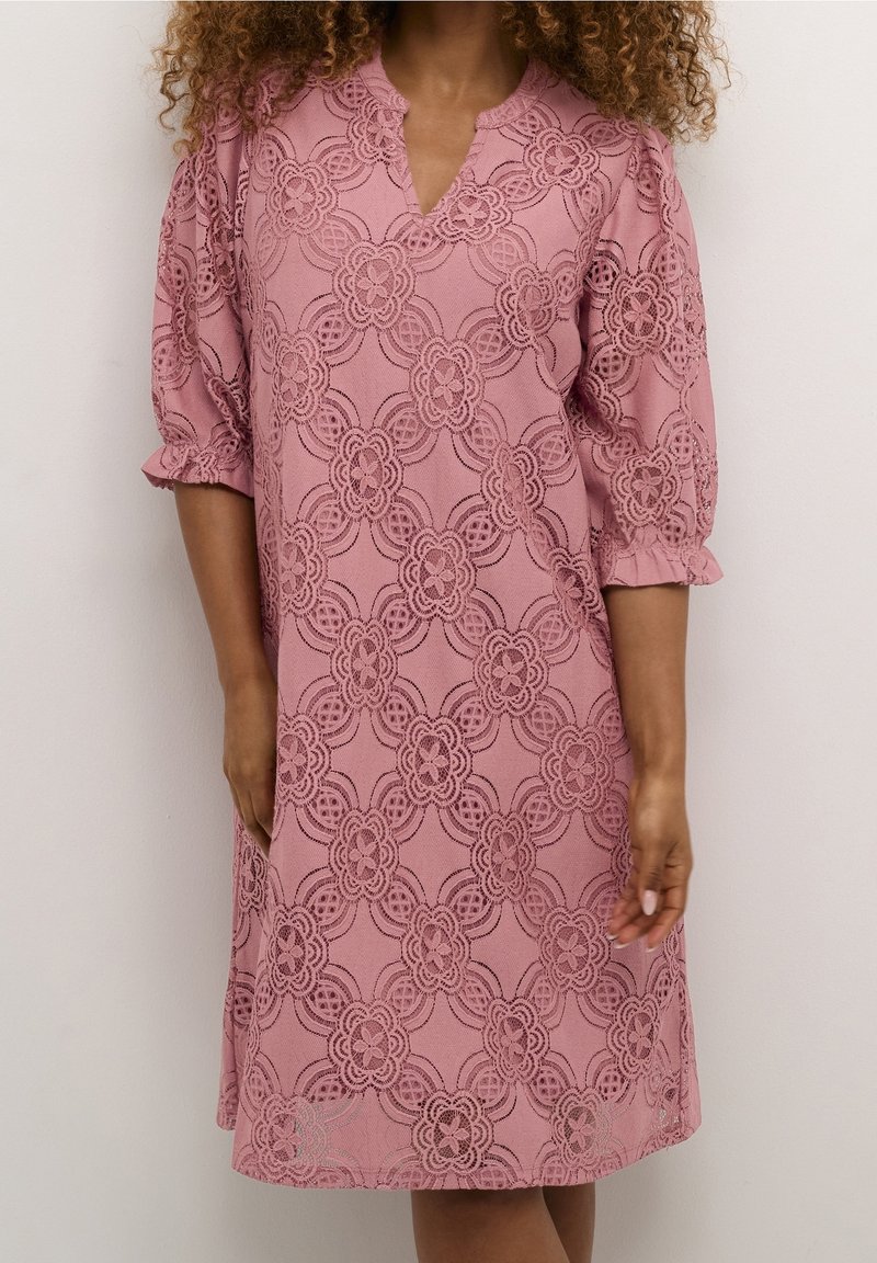 Femme portant une robe rose poudré longueur genou avec un motif en dentelle florale et des manches trois-quarts bouffantes, debout devant un fond uni.