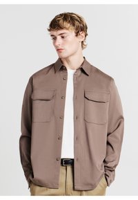 Braunes Hemd mit Knopfleiste und Kragen, zwei Brusttaschen und glatter Textur, kombiniert mit einem hellgrauen Unterhemd und beigen Hosen.