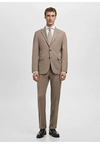 Jasnobrązowy garnitur z jednorzędową marynarką, klapami z wycięciem oraz dopasowanymi spodniami. Uzupełniony białą koszulą i jasnym krawatem.