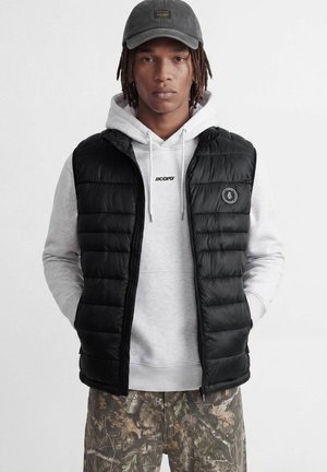 Jeune homme portant une casquette noire, un gilet matelassé noir, un sweat à capuche gris clair et un pantalon camouflage, debout les mains dans les poches devant un fond blanc.