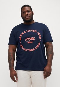 T-shirt in cotone blu navy con una grafica circolare che presenta "JACK&JONES 1990", "URBN." e "COLLECTORS CREW" in rosa. Maniche corte e colletto rotondo.