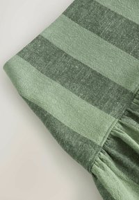 Tissu vert avec de larges rayures horizontales et des détails froncés près d'une couture sur un article textile, présenté sur un fond uni.