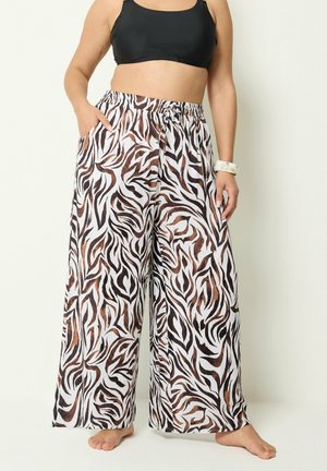 Kurvet person iført vidde-ben zebra-print bukser med elastisk talje og sort crop top, stående barfodet på en ensfarvet baggrund.