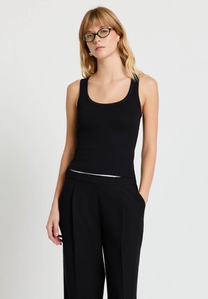 Giovane donna con occhiali che indossa una canotta nera senza maniche e pantaloni neri larghi, in piedi contro uno sfondo semplice.