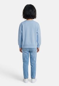 Enfant aux cheveux noirs courts portant un sweat-shirt bleu clair, un jean bleu clair et des baskets blanches, debout de dos sur un fond blanc.