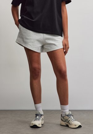 Person iført lysegrå shorts, en sort t-shirt, hvide sokker og hvide og grå løbesko, stående på en grå gulv.
