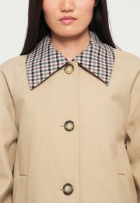 Cappotto beige con un ampio colletto a scacchi in tonalità bianco e nero. Presenta bottoni rotondi e una texture liscia e morbida.