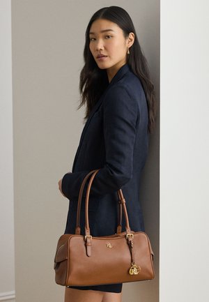 Kvinde i mørkeblå blazer, der holder en brun lædertaske med RL-logo og guldtaskevedhæng, stående foran en neutral væg.