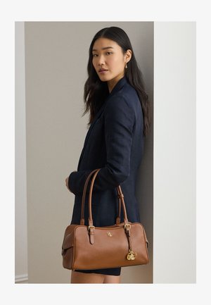 Kvinde i mørkeblå blazer, der holder en brun lædertaske med RL-logo og guldtaskevedhæng, stående foran en neutral væg.