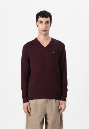 Pull en V bordeaux avec manches longues, bords-côtes aux poignets et à l'ourlet, et petit logo sur la poitrine. Matière tricotée douce avec une texture lisse.