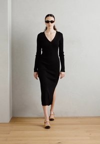Black ribbed midi dress with long sleeves, V-neck, and side slit, paired with modern yellow heels and dark sunglasses. Minimalist design.