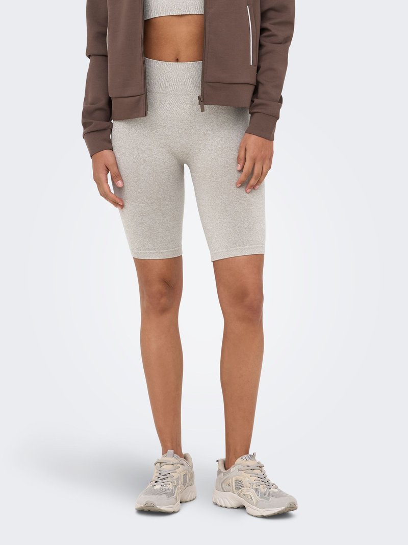 Personne portant un short de cycliste beige taille haute, un soutien-gorge de sport assorti, une veste zippée marron et des baskets de sport beiges, debout sur un fond uni.