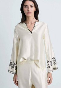 Cream satin blouse with a V-neckline, long sleeves, and black embroidered accents on cuffs. Smooth texture, relaxed fit.