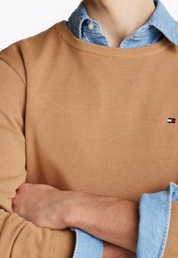 Homme portant une chemise en jean bleu clair sous un pull de couleur camel avec un petit logo rouge, blanc et bleu, bras croisés sur la poitrine.