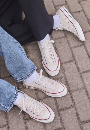 Deux paires de baskets en toile beige avec des lacets blancs portées avec des chaussettes blanches, l'une portée avec un jean bleu et l'autre avec un pantalon noir, sur des pavés.