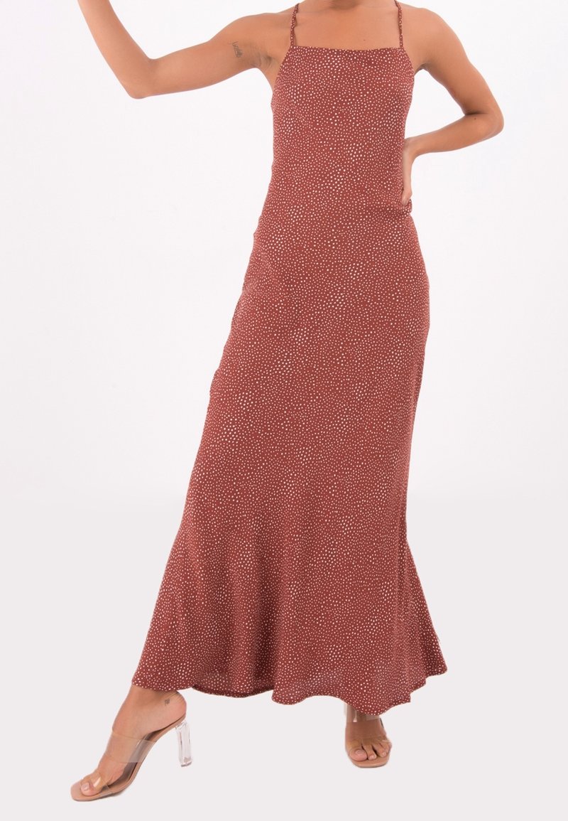 Femme portant une robe longue couleur rouille à petits pois blancs avec fines bretelles, associée à des sandales à talons transparents.