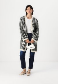 Cardigan com padrão preto e branco, de corte oversized, textura de malha, com frente aberta, usado por cima de uma blusa branca, combinado com jeans azuis e sapatos claros.