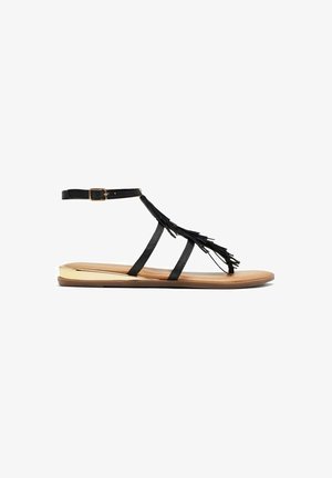 Czarne skórzane sandały gladiatorki z frędzlami, regulowanym paskiem na kostkę i płaską, brązową podeszwą z metalicznym akcentem.