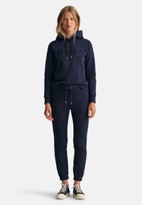 Mørkeblå hettegenser med matchende joggebukser. Laget av myk stoff, har logo trykk på forsiden og elastiske mansjetter med snor.