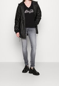 Chaqueta negra ligera con un acabado texturizado, top negro de escote en pico con detalles de pedrería, jeans ajustados grises y zapatos de tacón grueso negros.