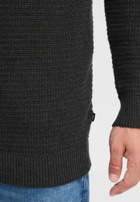 Schwarzer, strukturierter Pullover mit gerippten Bündchen und Saum. Verfügt über ein gewebtes Muster und ein kleines Etikett an der Seite. Eine Hand ist am unteren Rand sichtbar.