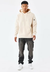 Sudadera con capucha beige, con bolsillo frontal, cordones y detalle en la manga izquierda, combinada con jeans de mezclilla oscura y zapatillas blancas.