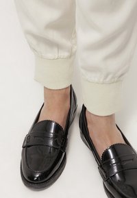 Zwarte gepatineerde leren instappers met een gestikt ontwerp en decoratieve hardware, gecombineerd met crèmekleurige elastische broek met manchet.